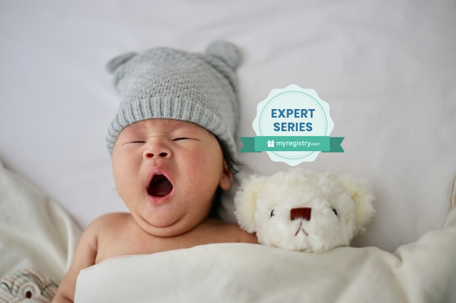When should your baby sleep through the night?, a yawning baby lying on a bed, under the covers, with a white plush bear tucked in beside him.